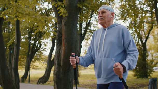 Aktiivinen vanhempi valkoihoinen mies isoisä käyttää kuulokkeita harjoitellaan Nordic kävely askel hiihto vaellus pylväät, kuunnella musiikkia puistossa. Retkeilyharrastus. Eläkkeellä olevat vanhukset terve urheilu - Valokuva, kuva