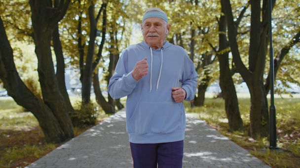 Atletyczna kondycja seniora starszego biegacza sportowego trening człowieka, słuchanie muzyki w słuchawkach. Trening cardio na zewnątrz w parku rano. Dziadek cieszy się zdrowym trybem życia. Aktywni emeryci dojrzali - Zdjęcie, obraz