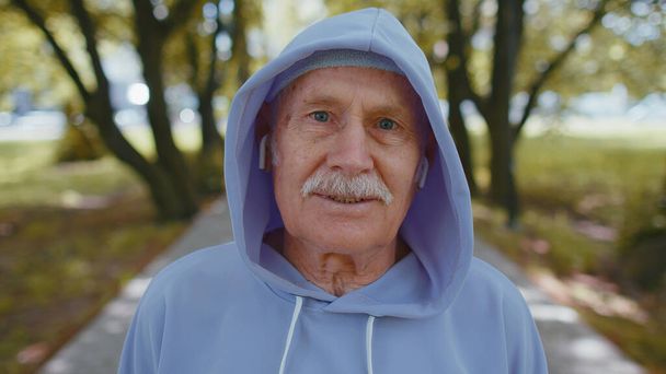 Portrait of athletic fitness senior elderly sport runner man training, listening music in earphones, wearing hood, looking at camera and smiling. Grandfather workout cardio outside in park at morning - Photo, Image