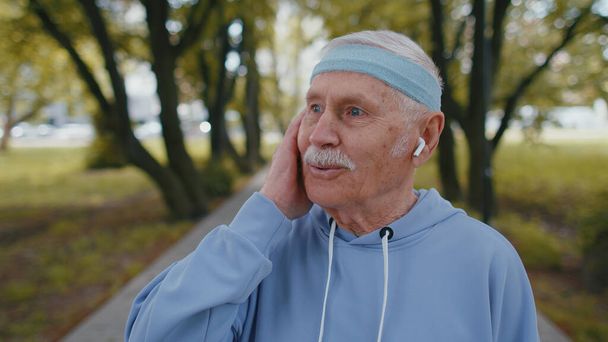 Portret sportowca starszego starszego biegacza sportowego, noszącego słuchawki słuchające muzyki, patrzącego w kamerę i uśmiechającego się. Zdrowy dziadek trening cardio na zewnątrz w parku w godzinach porannych - Zdjęcie, obraz