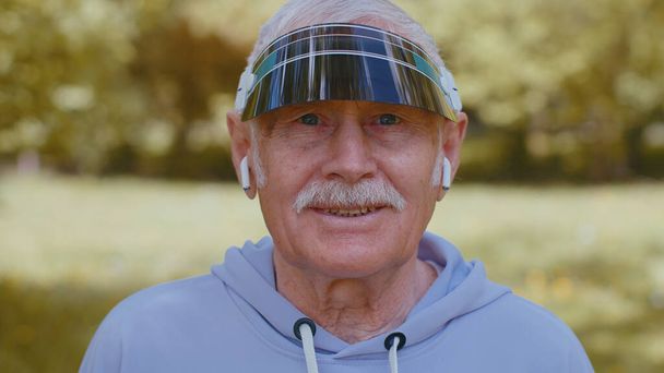 Portret sportowca starszego starszego biegacza sportowego, noszącego słuchawki słuchające muzyki, patrzącego w kamerę i uśmiechającego się. Zdrowy dziadek trening cardio na zewnątrz w parku w godzinach porannych - Zdjęcie, obraz