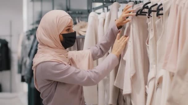Jonge geconcentreerde arabische vrouw shopper in medisch masker dragen hijab kiest bovenkleding in kleding winkel gelukkig meisje koper vindt geschikte jas tevreden klant vreugde met verkoop uitstekende korting - Video