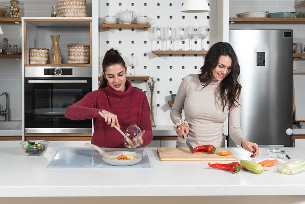 Two young happy college student roommates or business women cooking food together at their apartment. Females having fun together preparing meal in their modern kitchen at cozy home. - Φωτογραφία, εικόνα