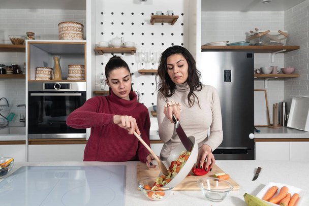 Two young happy college student roommates or business women cooking food together at their apartment. Females having fun together preparing meal in their modern kitchen at cozy home. - Fotoğraf, Görsel
