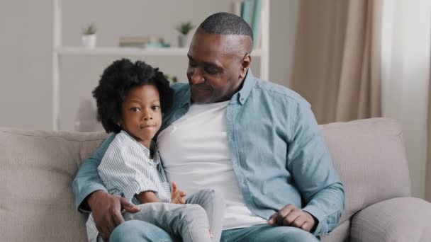 Šťastný africký americký rodinný dospělý otec s malou legrační dcera sedí na pohovce v pokoji s úsměvem tráví čas společně při pohledu na kameru mávání pozdravy nahrávání vlog muž mluví na webkameru - Záběry, video