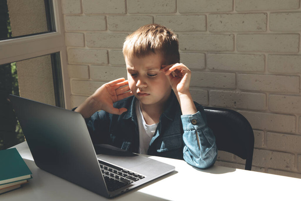 Schoolboy est assis au bureau en utilisant un ordinateur portable gris près de la fenêtre - Photo, image
