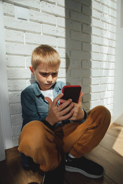 Boy 6-7 years old in a blue shirt sitting on the floor near the window carefully looks into the smartphone. - Zdjęcie, obraz