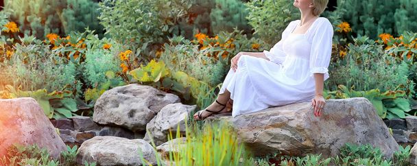 Banner with a woman dressed in a white dress sitting on a stone. Background - nature, park. . Concept nature and beauty. Copy space on the left - 写真・画像
