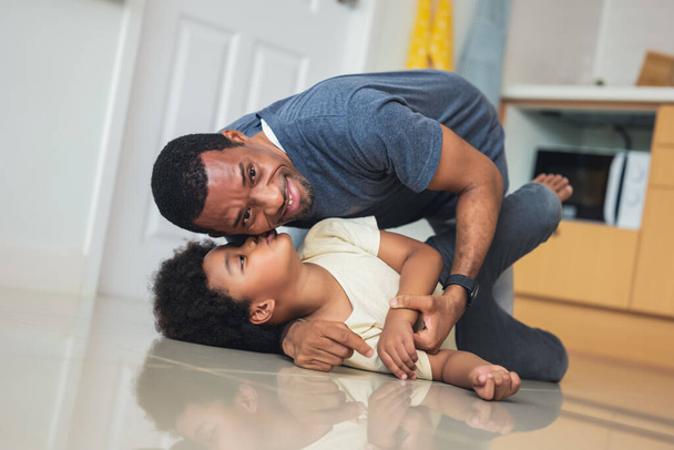 Musta perhe viettää aikaa yhdessä kotona. Hymyilevä afroamerikkalainen isä suutelee ja syleilee ihanaa poikaansa makaamassa lattialla keittiössä. Isyyden ihanat hetket käsite - Valokuva, kuva