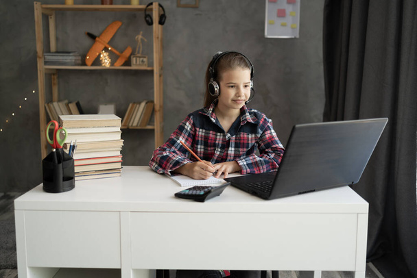 Ev ödevi yapan liseli kız dizüstü bilgisayar ve kulaklık kullanıyor, bir deftere notlar yazıyor zoom 'da sanal bir web sitesi dinliyor, e-öğreniyor, evde eğitim veriyor. - Fotoğraf, Görsel