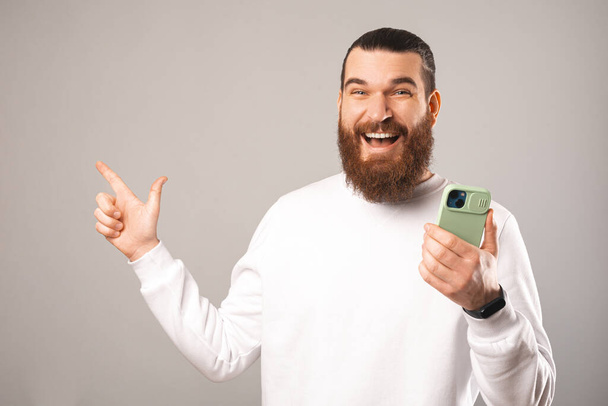 portrait of cheerful handsome young man holding new smarpthone and pointing away at copyspace over grey background. - Foto, imagen