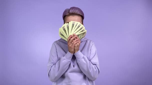 Modische junge erwachsene hübsche Frau in lila Kapuzenpulli mit kurzer Frisur versteckt sich hinter einem Bündel Banknoten, senkt das Geld, blickt vereinzelt lächelnd in die Kamera auf violettem Hintergrund drinnen - Filmmaterial, Video