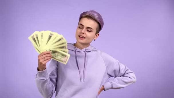 Mujer adulta joven afortunada con pelo púrpura corto sosteniendo un montón de dinero, ondeando billetes para enfrentar, mostrando satisfacción, disfrutando de una vida rica aislada en un fondo liso violeta en el interior. Riqueza, ganador - Metraje, vídeo