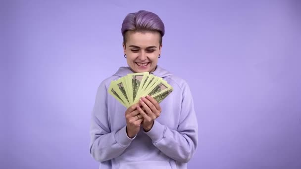 Πορτρέτο κατάπληκτος χαρούμενη σύντομη μωβ μαλλιά γυναίκα σε αστικό στυλ hoodie χαρά τύχη λαχείο, που δείχνει δέσμη των χαρτονομισμάτων δολάριο, υπερηφανεύεται για το κέρδος χρήματα. Εσωτερική στούντιο πυροβόλησε απομονώνονται σε βιολετί φόντο - Πλάνα, βίντεο