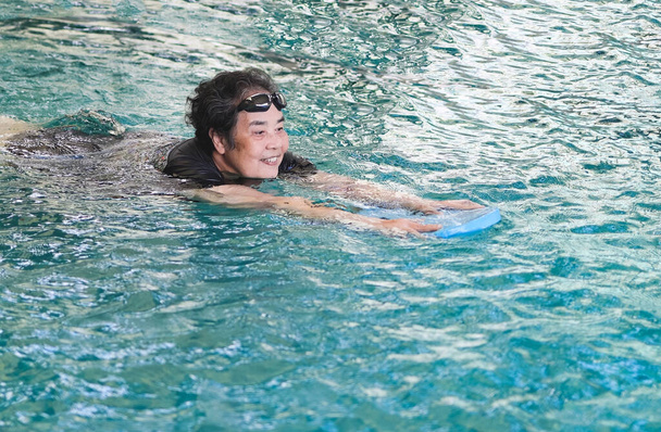 Portrait of  healthy Asian senior woman with kickboard in a swimming pool. Old woman swimming in water with the help of a kickboard. Smiling old woman swimming with inflatable board in swimming pool. - Photo, Image