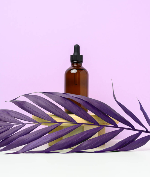 Ruskea lasipullo, jossa on pipetti puisessa korokkeessa ja violetteja palmunlehtiä. Kosmeettisten valmisteiden, öljyjen ja seerumien säilytyspäällykset - Valokuva, kuva