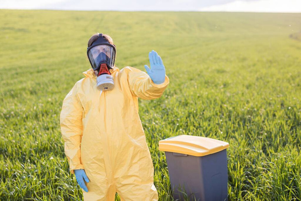 A woman in a yellow protective suit stands in the middle of a green field in a protective gas mask and shows a gesture to call to stop and save the planet. A woman stands near the garbage can and calls to save the planet - 写真・画像