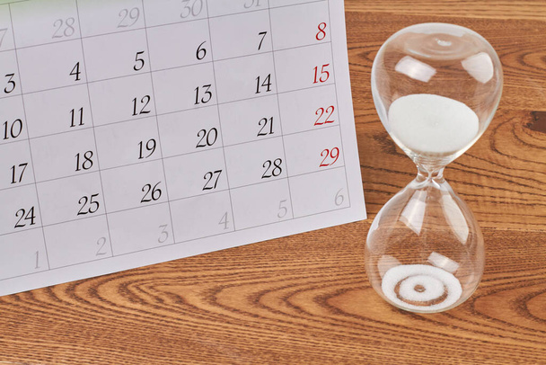 Close-up κλεψύδρα με λευκή άμμο και ημερολόγιο στο ξύλινο γραφείο. Χρονόμετρο αντίστροφης μέτρησης γυαλιού. - Φωτογραφία, εικόνα