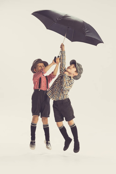 Portrait of two active little boys, children playing together with umbrella, jumping isolated over grey studio background. Concept of childhood, friendship, family, fun, lifestyle, retro fashion - Photo, image