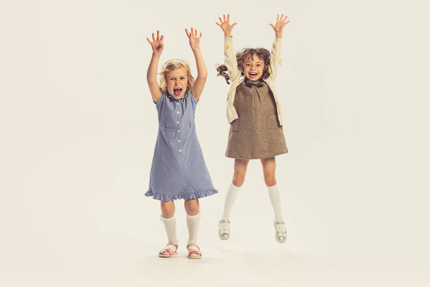 Портрет двух веселых маленьких девочек, прыгающих детей, играющих вместе на сером фоне студии. Мбаппе детства, дружбы, семьи, развлечений, образа жизни, ретро-моды - Фото, изображение