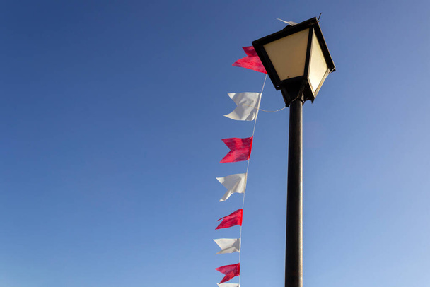 Una lámpara de calle vintage, con una línea de banderas blancas y rojas, con el cielo azul en el fondo. Poste de luz. Lámpara con banderas. - Foto, Imagen