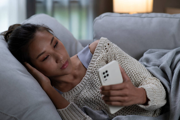 Frusztrált és depressziós fiatal ázsiai nő sír egy okostelefonnal a kezében, miközben ő fekszik a kanapén otthon, szomorú ázsiai nő kanapén érzi le olvasni rossz hír a mobiltelefon online. - Fotó, kép