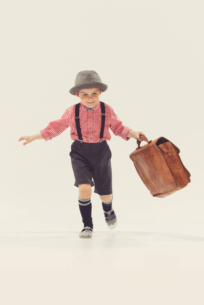 Portrait of little boy. child in checkered shirt with suitcase walking on tiptoe isolated over grey studio background. Concept of childhood, friendship, family, fun, lifestyle, retro fashion - Fotoğraf, Görsel