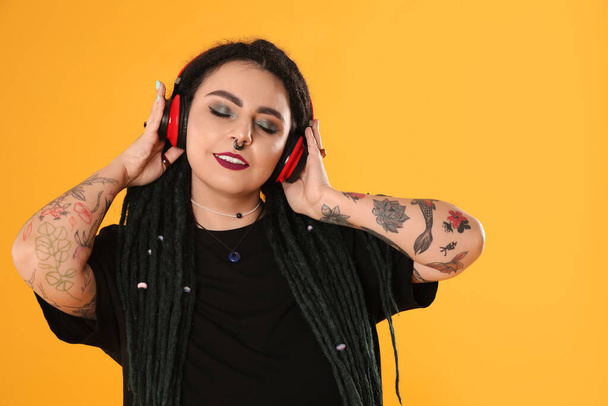 Beautiful young woman with tattoos on arms, nose piercing and dreadlocks listening music against yellow background - Foto, Imagen