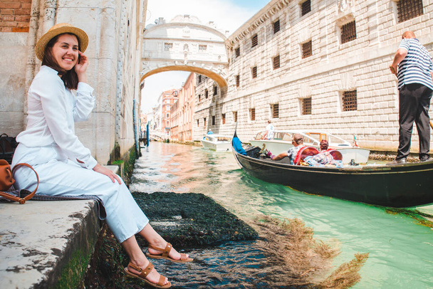 женщина, сидящая на городской набережной в Венеции, наслаждается видом на каналы с гондолами
 - Фото, изображение