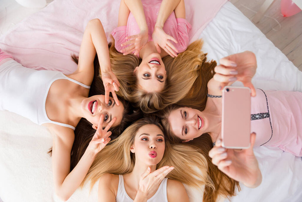 High angle view portrait of four girls laying white sheets make selfie show v-sign send air kiss indoors. - Foto, Bild