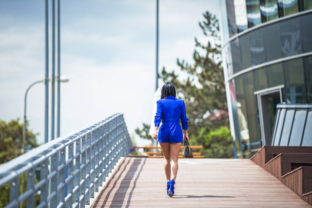 the woman is photographed from behind. The woman walks down the ramp towards the building and is dressed in a blue costume. The woman is in blue shoes and carries a brown handbag. - Photo, Image