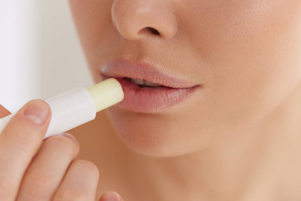 Lips care. Close up of young woman applying hygienic balm on her plump lips in studio. Charming lady using professional cosmetic for taking care of her soft skin. - Photo, Image
