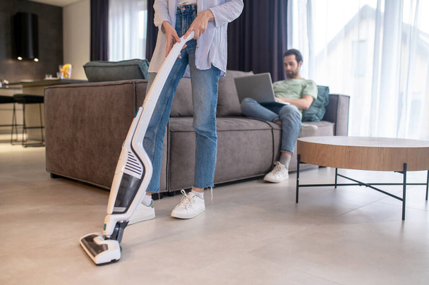 Vacuuming floor. Young bearded man reclining on sofa looking at laptop and woman vacuuming floor in modern apartment - Fotografie, Obrázek