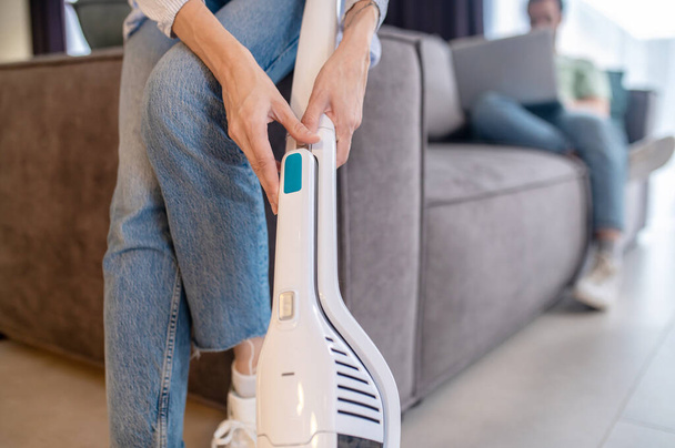 Ease of use. Female graceful hands touching modern vacuum cleaner near sofa at home and man with laptop behind - 写真・画像