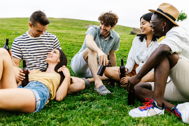 Ryhmä monirotuisia ystäviä hengailemassa ja puhumassa ulkona julkisessa puistossa Monipuoliset nuoret juomassa olutta yhdessä ja rentoutumassa ulkona kesällä - Valokuva, kuva