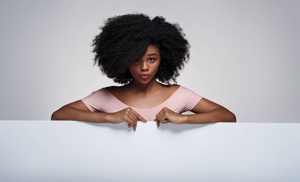 African American woman with curly hair looking at camera and pouting lips while pointing at blank poster during advertisement campaign against gray background - Fotó, kép
