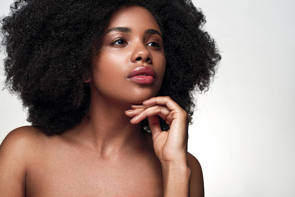 Pensive African American female model with bare shoulders and curly hair touching chin and looking away during skin care routine against gray background - Foto, Bild