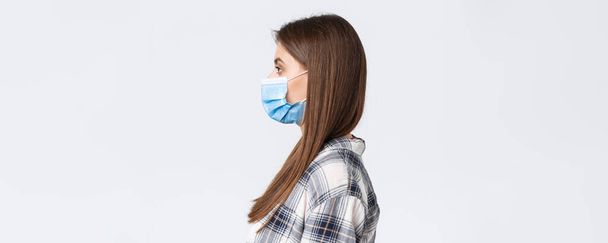 Coronavirus outbreak, leisure on quarantine, social distancing and emotions concept. Profile of serious-looking young pretty woman in medical mask standing in line, white background. - Photo, Image
