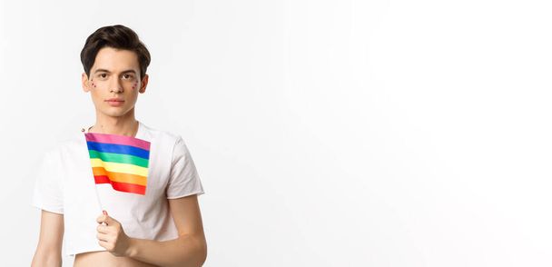 Pride and lgbtq concept. Waist up shot of attractive anrogynous man holding rainbow flag, having glitter on face and looking at camera, standing over white background. - 写真・画像