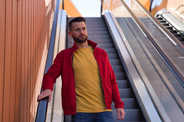Νέος όμορφος άντρας με κόκκινο μπουφάν, κίτρινο μπλουζάκι και τζιν κατεβαίνει κυλιόμενες σκάλες σε ένα εμπορικό κέντρο. Concept ομορφιά, μόδα, μοντέρνα, μοντέρνα. - Φωτογραφία, εικόνα
