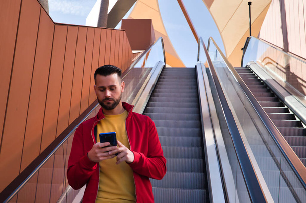 Handsome young man in red jacket, yellow t-shirt and jeans checking social networks on his cell phone while going down escalators in a shopping mall. Smartphone concept, fashion, modern, trendy. - Fotoğraf, Görsel
