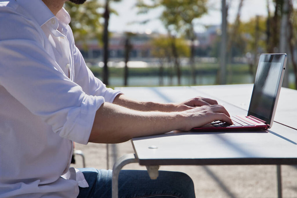 Detalle de manos y brazos del joven, en camisa blanca, trabajando en la calle con su portátil sobre una mesa de madera blanca. Concepto de tecnología, trabajo remoto, netbook, tablet. - Foto, Imagen