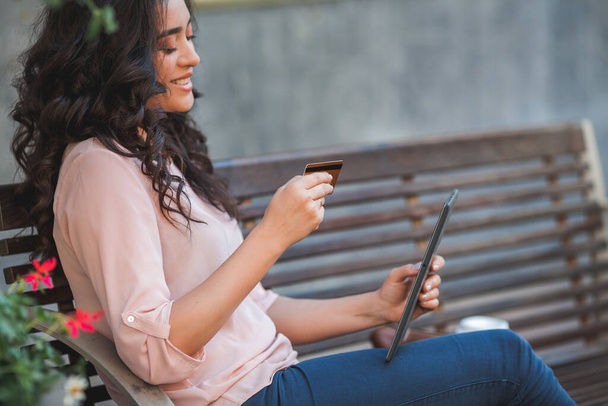 Młoda ciemnowłosa kręcona kobieta płaci za zakupy przez internet za pomocą tabletu. 35-letnia modelka siedzi na ulicy w centrum miasta. - Zdjęcie, obraz