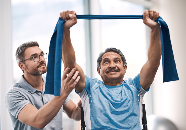 Aufnahme eines älteren Mannes beim Training mit einem Widerstandsband während einer Rehabilitationseinheit mit seinem Physiotherapeuten. - Foto, Bild