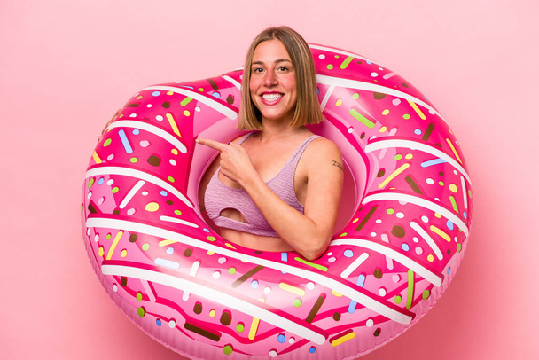 Joven mujer caucásica sosteniendo un colchón de aire aislado sobre fondo rosa sonriendo y señalando a un lado, mostrando algo en espacio en blanco. - Foto, Imagen