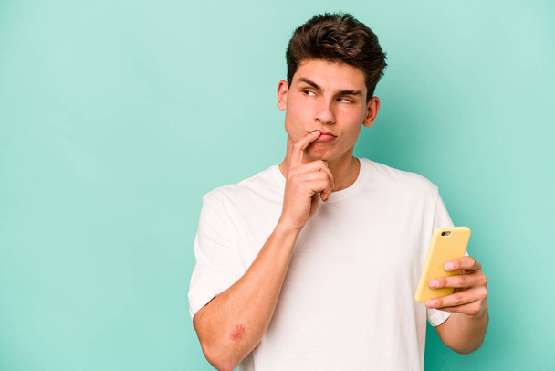 Jeune homme caucasien tenant téléphone portable isolé sur fond bleu détendu penser à quelque chose qui regarde un espace de copie. - Photo, image