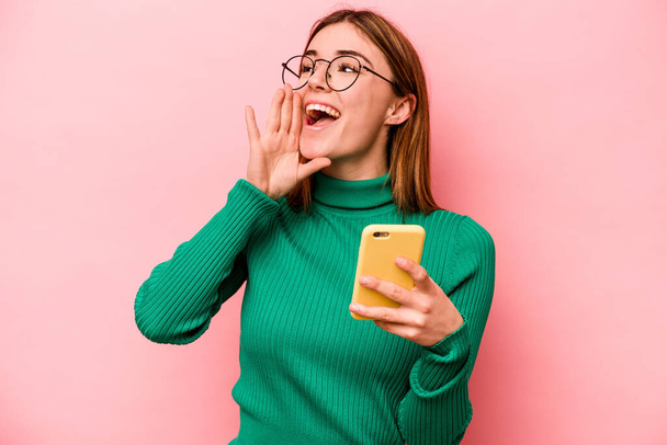 Giovane donna caucasica in possesso di cellulare isolato su sfondo rosa gridando e tenendo palmo vicino alla bocca aperta. - Foto, immagini