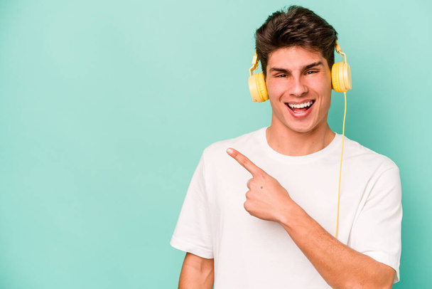 青い背景の上に隔離された音楽を聴いている若い白人男性は笑って脇を指差して、空白のスペースで何かを示す. - 写真・画像