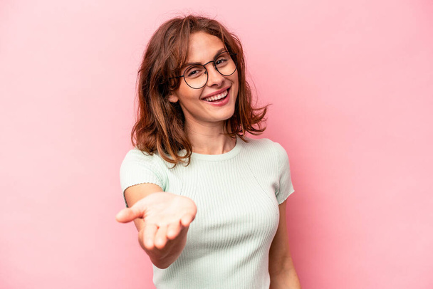 Young caucasian woman isolated on pink background stretching hand at camera in greeting gesture. - Foto, imagen