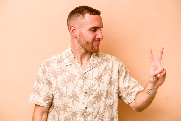 Young caucasian man isolated on beige background joyful and carefree showing a peace symbol with fingers. - Photo, Image